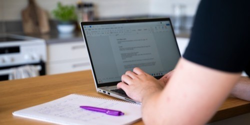 En person som sitter och skriver en text på en laptop med ett anteckningsblock bredvid.