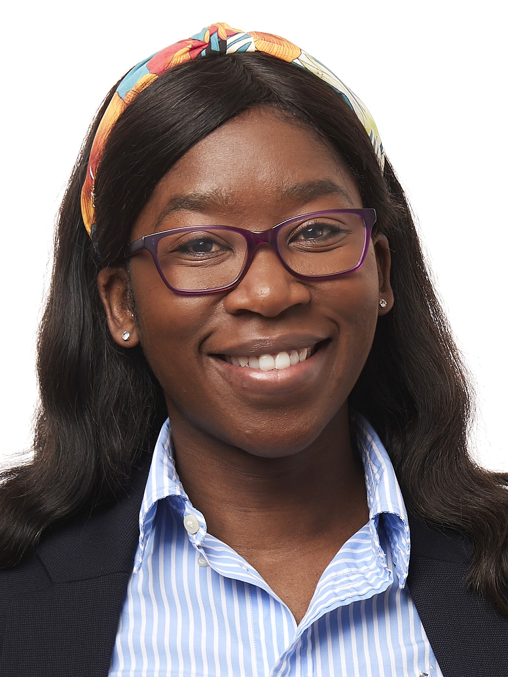Woman wearing glasses, a colorful headband, and a blue striped shirt under a dark blazer, smiling and facing forward against a white background.