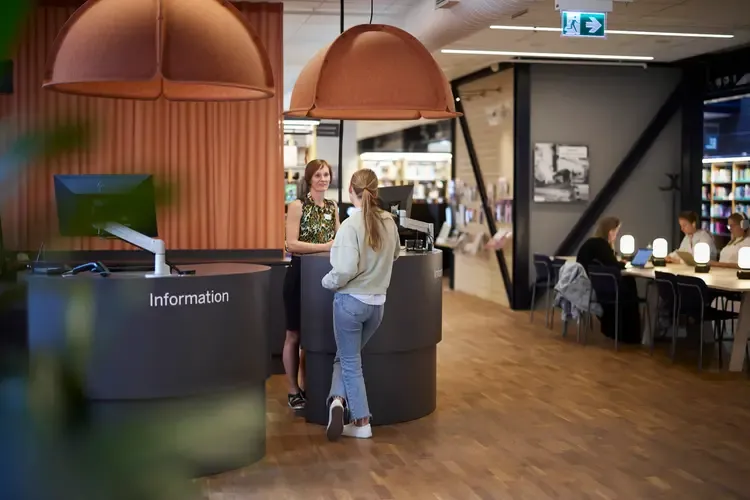 Bibliotekets informationsdisk, där det står en bibliotekarie och pratar med en student. Över disken hänger två stora lampor i en orange nyans. I bakgrunden syns ett bord där några studenter sitter och studerar.