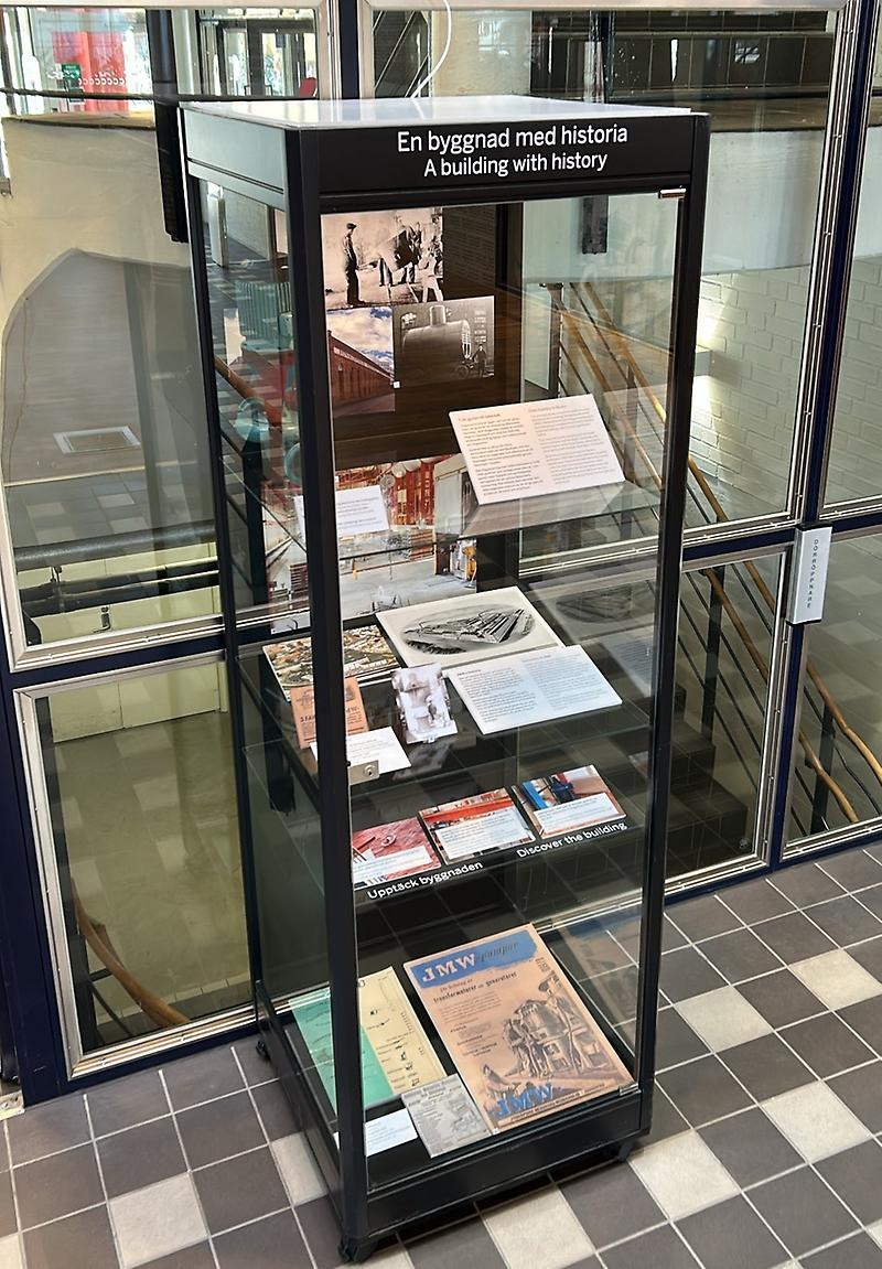 En svart glasmonter med olika bilder och prylar i. Montern står på ett grått och vitt rutigt golv.